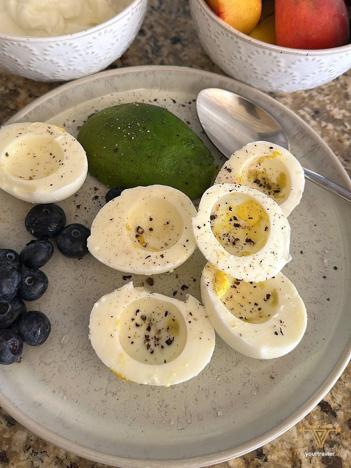 Halved boiled eggs with black pepper, half an avocado and blueberries on a grey plate