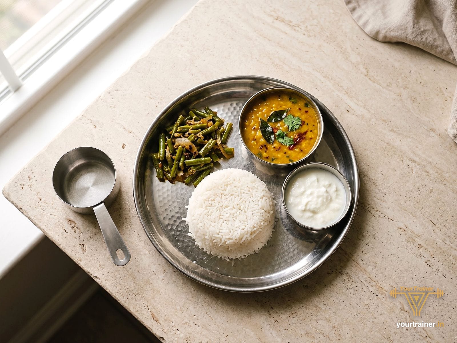 Steel thali holding a measured cup of white rice, a katori of dal, a katori of curd and green beans, with a measuring cup beside it