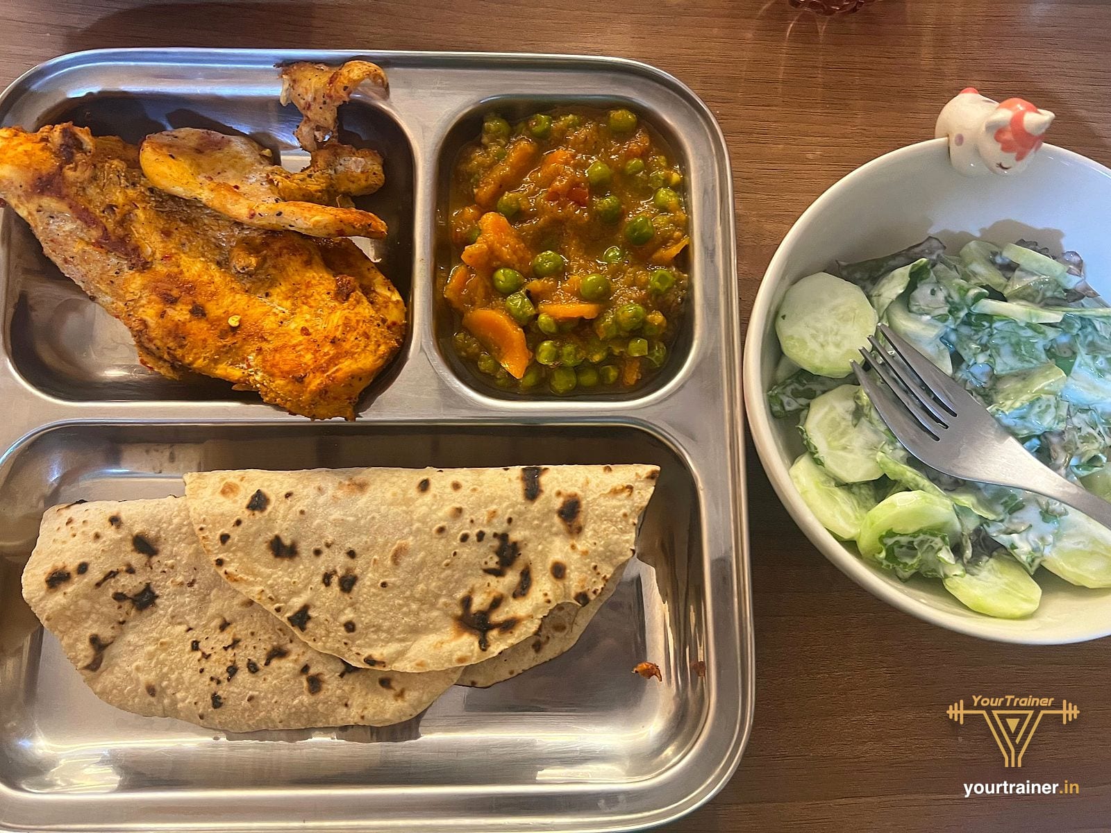 Steel tray with grilled chicken, two rotis and a peas and carrot sabzi, with a bowl of cucumber raita