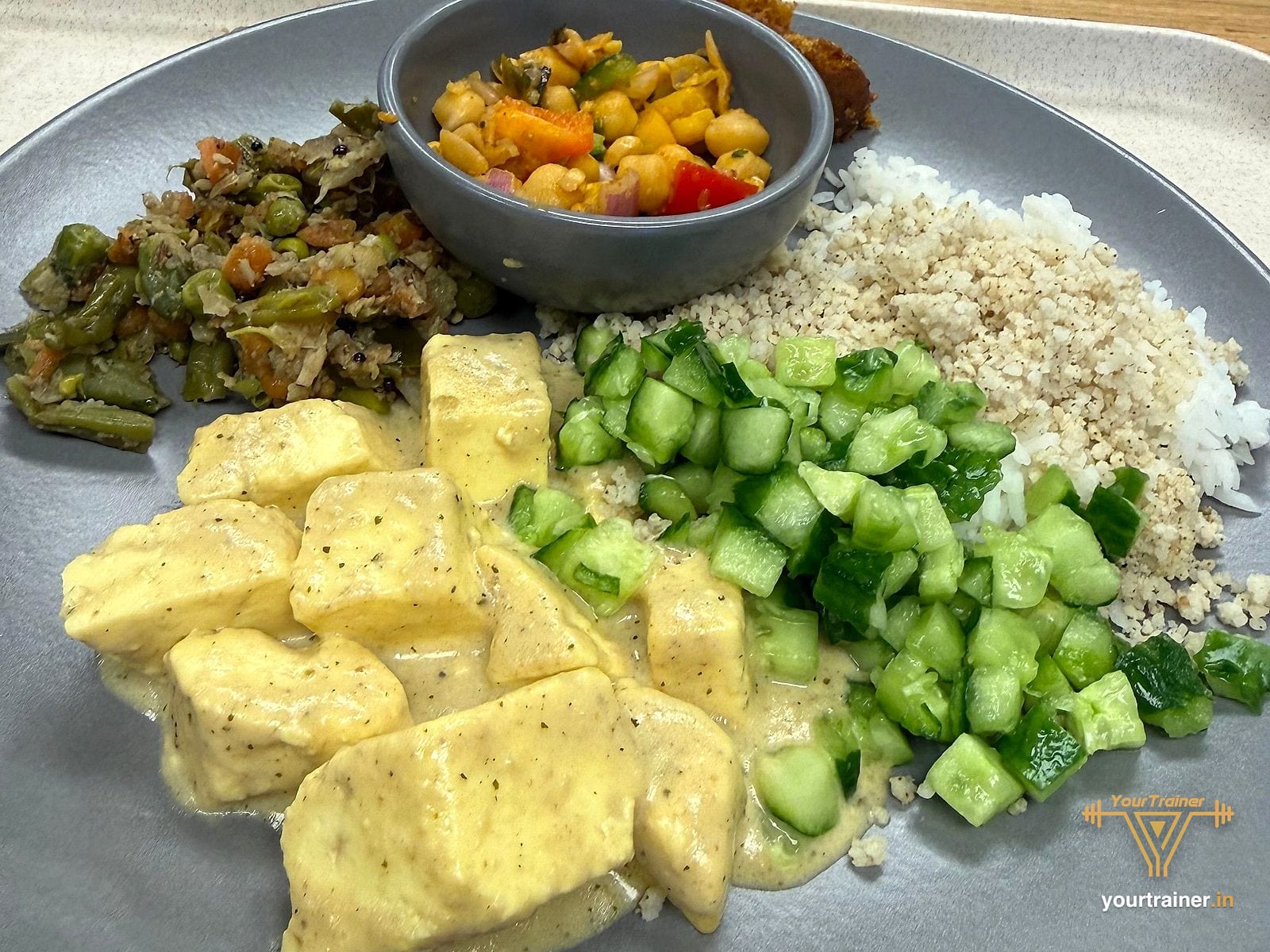 Plate of paneer in a light curry with millet and rice, chopped cucumber, beans sabzi and a bowl of chana salad